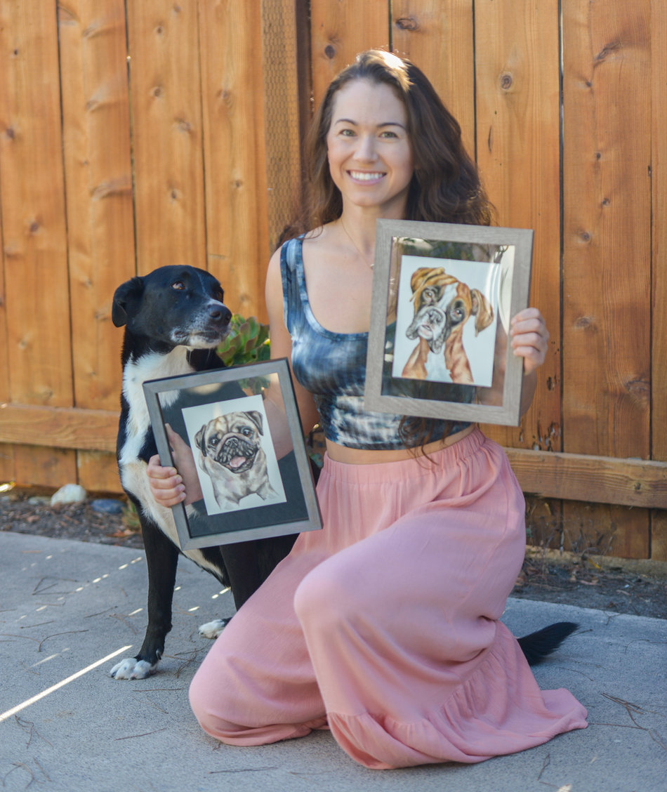 Lauren Salas holding two framed watercolor pet portraits, with her dog Roscoe