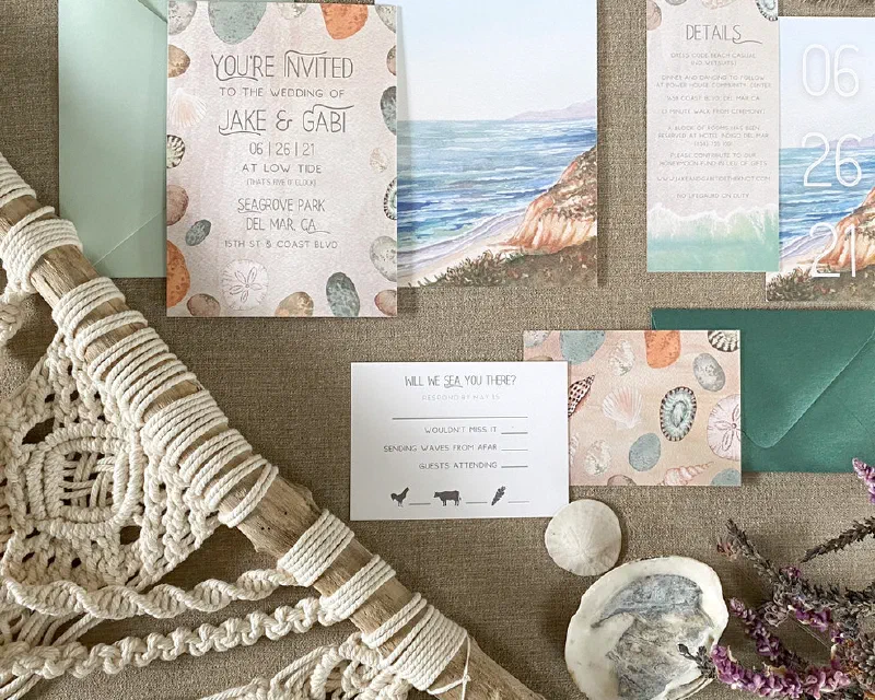 Coastal watercolor wedding invitation suite with a beach scene, shells, and a sage-green envelope