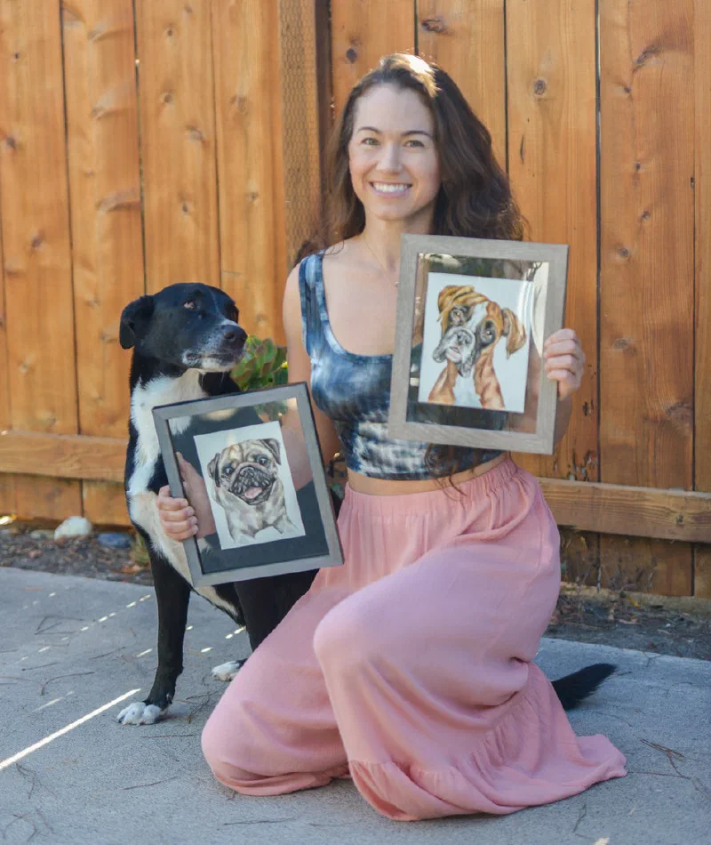 Lauren Salas holding two framed watercolor pet portraits, with her dog Roscoe
